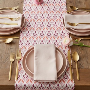 May include: A table setting with a floral patterned table runner in shades of pink and red. The setting includes pink plates, napkins, and gold-colored cutlery. Fresh flowers are placed on the table, enhancing the elegant dining arrangement.