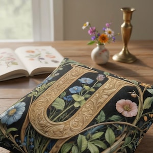 Op de afbeelding: Decoratief sierkussen met een zwarte achtergrond met een gouden letter "U" gevuld met bloemenillustraties in blauw, roze en groen. Het kussen ligt op een houten oppervlak met een boek en een vaas op de achtergrond.