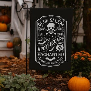 Peut inclure: Drapeau de jardin noir avec texte blanc "Olde Salem Apothecary", avec un crâne, des os croisés, des chauves-souris et un chaudron. Le drapeau est exposé à l'extérieur avec des citrouilles et des feuilles d'automne.