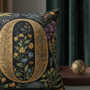 Op de afbeelding: Decoratief sierkussen met een donkere achtergrond en een grote, sierlijke gouden letter "O" gevuld met kleurrijke bloemen. Het kussen heeft een bloemenpatroon met gele, blauwe en paarse bloemen en groene bladeren. Het kussen is vierkant.
