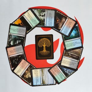 Puede incluir: Un conjunto de cartas de Magic: The Gathering dispuestas en círculo. Las cartas presentan ilustraciones de paisajes y texto. Una carta central muestra un diseño de árbol dorado sobre fondo negro. El conjunto se encuentra sobre un fondo rojo y blanco.