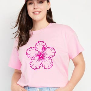 May include: A light pink t-shirt featuring a large, magenta hibiscus flower graphic. The flower is centered on the shirt and has detailed petals and a prominent pistil. The shirt is made of a soft material.