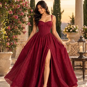 Può includere: Un abito lungo color bordeaux intenso caratterizzato da un corpetto strutturato in stile corsetto con spalline sottili, scollo a cuore e un'ampia gonna voluminosa con un alto spacco sulla coscia. Il tessuto presenta un motivo a foglie tonale.