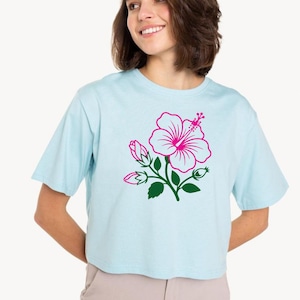 May include: Light blue crop top featuring a pink hibiscus flower design with green leaves and buds. The floral graphic is centered on the front of the shirt. The shirt has short sleeves and a crew neck.