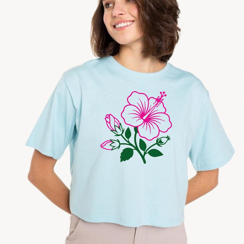 May include: Light blue crop top featuring a pink hibiscus flower design with green leaves and buds. The floral graphic is centered on the front of the shirt. The shirt has short sleeves and a crew neck.