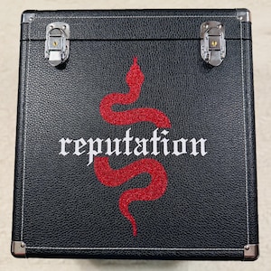 Pode incluir: Caixa preta com ferragens prateadas e um design de cobra vermelha com glitter. A palavra "reputation" está escrita em branco com fonte gótica abaixo da cobra. A caixa tem uma superfície texturizada e costuras brancas.