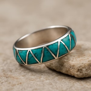 Op de afbeelding: Een zilveren ring met een turkoois inleg. Het turkoois is in driehoekige vormen gesneden en gezet in een zilveren band. De ring wordt getoond op een lichtbruine steen.