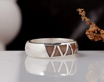 Op de afbeelding: Een zilveren ring met een band met een reeks bruine driehoekige inlegwerk. De ring heeft een gepolijste afwerking en een eenvoudig, elegant ontwerp. De ring wordt gepresenteerd op een wit oppervlak met een donkere achtergrond.