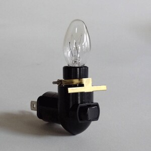 May include: A black night light with a clear, teardrop-shaped bulb. The night light has a brass-colored metal bracket and a small on/off switch. The plug is a two-prong type.