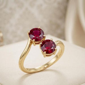 Può includere: Un anello aperto in oro giallo lucido, caratterizzato da due rubini rossi intensi a taglio rotondo montati su quattro griffe. Il design aperto permette alle due pietre di posizionarsi fianco a fianco, creando un aspetto distinto ed equilibrato.