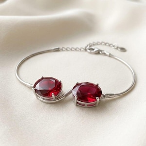 Op de afbeelding: Een zilverkleurige armband met twee ovale, dieprode gefacetteerde stenen in klauwzettingen. De armband is gemaakt van een flexibele slangenketting en bevat een verstelbare verlengketting met een kleine ronde bedel aan het uiteinde.