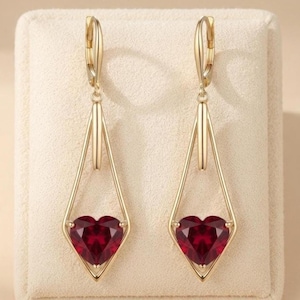 Puede incluir: Un par de pendientes colgantes de tono dorado con un marco geométrico en forma de diamante. Cada pendiente suspende una piedra preciosa roja oscura en forma de corazón facetado en la base, asegurada por un cierre de palanca articulado.