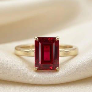 Puede incluir: Un anillo de oro con una gran piedra preciosa roja de corte esmeralda rectangular, montada en cuatro garras. La banda es lisa y pulida, mostrando un diseño limpio que resalta el color rojo vibrante y profundo de la piedra central.