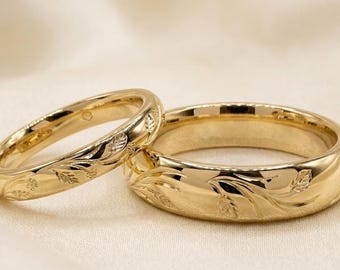 May include: Two polished yellow gold rings featuring an engraved leaf and floral pattern along the outer band. The rings have a smooth, reflective finish and a rounded profile, showcasing intricate botanical details etched into the metal surface.