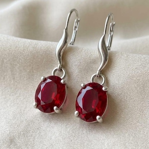 Puede incluir: Un par de pendientes colgantes de tono plateado con piedras facetadas de color rojo intenso de corte ovalado, sujetas por engastes de cuatro garras. Los pendientes tienen un tallo metálico curvo y ondulado que une la piedra al cierre.