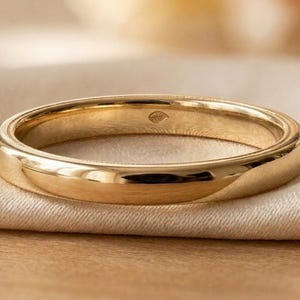 Può includere: Un anello a fascia in oro lucido con una superficie liscia e arrotondata e una finitura molto brillante. L'interno della fascia presenta una piccola incisione a forma di foglia, visibile lungo la circonferenza interna dell'anello in metallo.