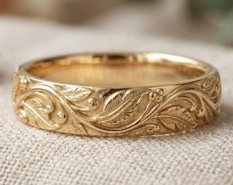 May include: A gold band ring featuring an intricate, continuous relief pattern of engraved leaves, vines, and small berries wrapping around the entire exterior surface of the metal, showcasing a polished finish with detailed textures.