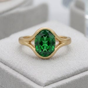 Puede incluir: Un anillo de oro con una gran piedra preciosa de color verde vibrante y corte ovalado, engastada en una banda de metal liso y pulido. El engaste presenta un diseño de vástago dividido que se curva alrededor de la piedra central.