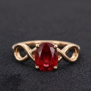 May include: A gold ring with a large, oval-cut, deep red gemstone. The ring's band has an infinity symbol design on either side of the gemstone. The gemstone is held in place by four prongs, creating a secure setting.