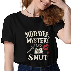 Può includere: T-shirt nero con la scritta bianca "MURDER, MYSTERY, AND SMUT". Il design include un coltello, un libro aperto e labbra rosse. La maglietta è abbinata a jeans blu scuro.