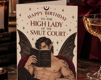 May include: A greeting card featuring an illustration of a shirtless, winged figure with pointed ears sitting in a chair and reading a book. The text reads Happy Birthday to the High Lady of the Smut Court, and the book cover says Good Girls Read Spicy Books.