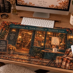 May include: A rectangular desk mat featuring a detailed illustration of a cosy bookstore exterior in autumn. The scene includes a storefront with shelves of books, warm lighting, fallen leaves, a small table with a mug, and text reading Good Books, Warm Drinks, Brighter Days, and Just One More Chapter.