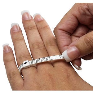 May include: A white ring sizer is wrapped around a finger to measure its size. The sizer has black numbers and markings, with the arrow pointing to the size. The finger is on a light skin tone.