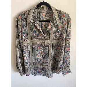 May include: A long-sleeved button-down shirt featuring a repeating grid pattern of floral motifs in shades of muted blue, beige, and red. The garment has a pointed collar, a front button placket, and buttoned cuffs, showcasing a structured geometric layout.