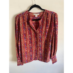 May include: A long-sleeved blouse with a red background and a vertical floral pattern in blue, yellow, and orange. The blouse has a V-neck and buttoned cuffs. The blouse is hanging on a black hanger.