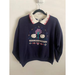 May include: Navy blue sweatshirt with a white collar featuring red heart accents. The front displays an embroidered design with a tree, clouds, and the text "COUNT YOUR BLESSINGS". A vintage-style, cozy top.