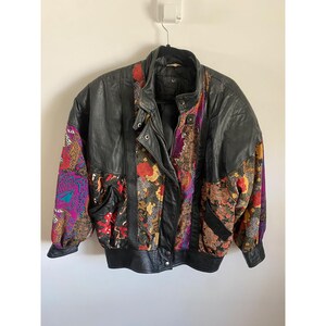 May include: A black leather jacket featuring panels of multi-colored fabric with floral and geometric patterns. The jacket includes a front zipper closure, snap buttons, long sleeves with ribbed cuffs, and a high collar. The design incorporates vibrant shades of red, purple, yellow, and orange throughout the patterned sections.