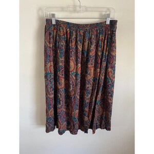May include: A knee-length skirt featuring an intricate paisley pattern in shades of deep red, golden yellow, and teal blue. The skirt has a gathered elastic waistband and hangs in soft, vertical folds from a plastic clothing hanger.