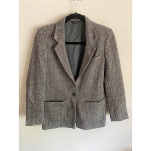 May include: A gray herringbone blazer with a single-button closure and two front pockets. The blazer is hanging on a black hanger against a neutral background. The interior lining is a dark gray color.