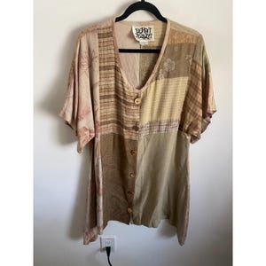 May include: A short-sleeve button-down shirt featuring a patchwork design in various shades of beige, tan, and brown. The garment has a V-neckline, five light-colored buttons, and a label at the neck that reads "Denny Jessi".