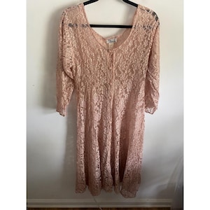 May include: A dusty rose lace dress featuring long sleeves, a V-neckline, and a front button closure. The garment is crafted from a floral patterned lace fabric and hangs from a black hanger, displaying a mid-calf length and a flared skirt.