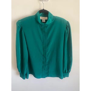 May include: A solid emerald green long-sleeve blouse featuring a high mock neckline, a concealed front button placket, and gathered puff sleeves with buttoned cuffs. The interior collar label displays the brand name Jordan in gold lettering.