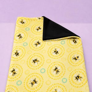May include: A yellow blanket with a repeating pattern of black and yellow bees inside of yellow circles. The blanket has a black backing. The blanket is rectangular and folded over.