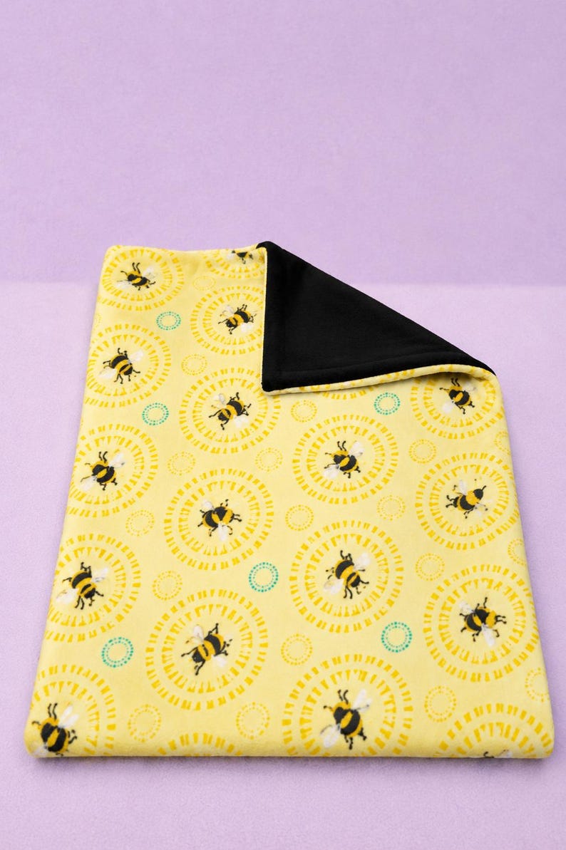 May include: A yellow blanket with a repeating pattern of black and yellow bees inside of yellow circles. The blanket has a black backing. The blanket is rectangular and folded over.