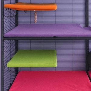 Puede incluir: Una jaula para mascotas de metal negro de varios niveles con cuatro plataformas rectangulares cubiertas con almohadillas de tela suave. Las almohadillas son de color naranja, morado, verde lima y rosa brillante. Un juguete de madera cuelga debajo.