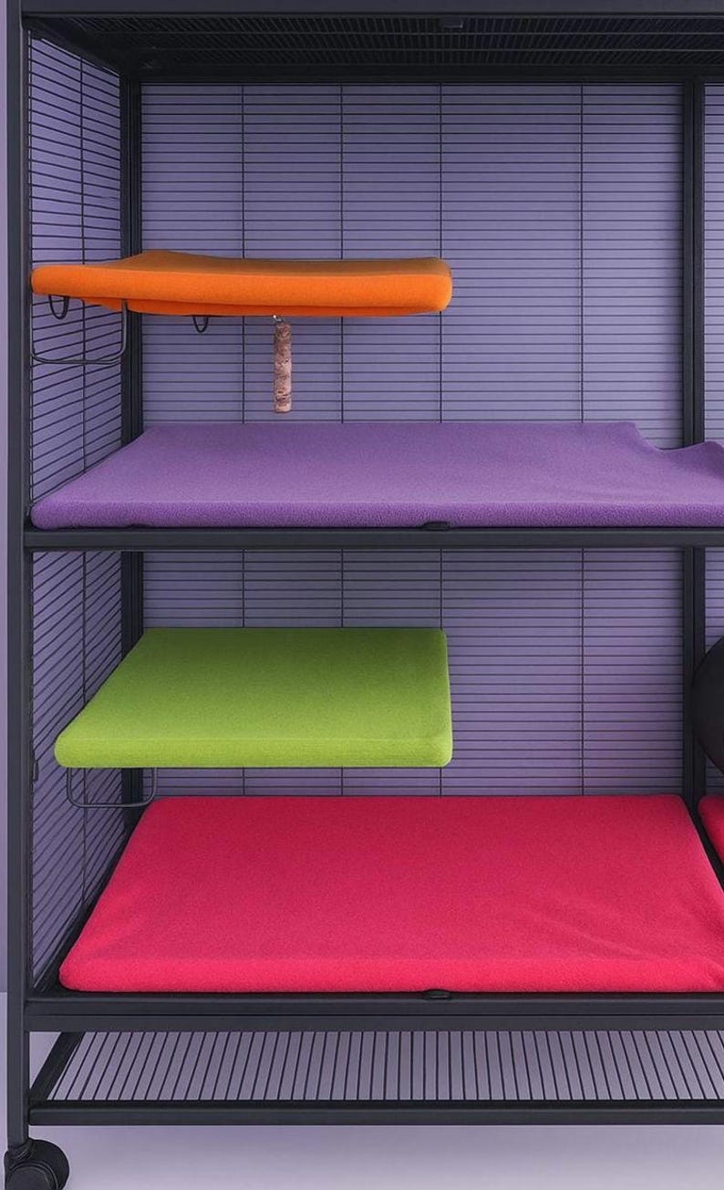 Puede incluir: Una jaula para mascotas de metal negro de varios niveles con cuatro plataformas rectangulares cubiertas con almohadillas de tela suave. Las almohadillas son de color naranja, morado, verde lima y rosa brillante. Un juguete de madera cuelga debajo.