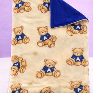 May include: A cream-colored baby blanket featuring a pattern of brown teddy bears wearing blue sweaters with the letter "A". The blanket has a solid blue border and is decorated with small gold stars.