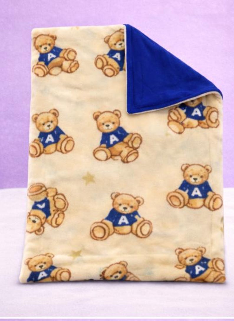 May include: A cream-colored baby blanket featuring a pattern of brown teddy bears wearing blue sweaters with the letter "A". The blanket has a solid blue border and is decorated with small gold stars.