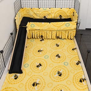 May include: A guinea pig cage with a yellow bee-themed bedding set. The set includes a patterned base, a black ramp, and a matching skirt. The cage is enclosed by a black wire frame.