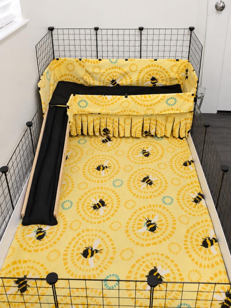 May include: A guinea pig cage with a yellow bee-themed bedding set. The set includes a patterned base, a black ramp, and a matching skirt. The cage is enclosed by a black wire frame.