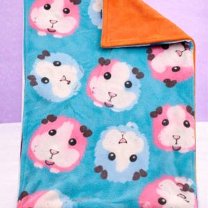 May include: A folded fleece blanket featuring a repeating pattern of cartoon guinea pig faces in pink and blue on a turquoise background. The reverse side is solid orange.
