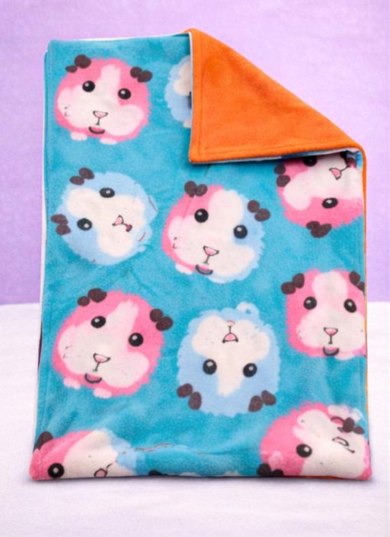 May include: A folded fleece blanket featuring a repeating pattern of cartoon guinea pig faces in pink and blue on a turquoise background. The reverse side is solid orange.