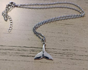Può includere: Una collana color argento con un ciondolo a forma di coda di balena. La catena è sottile e ha una chiusura regolabile. La collana è esposta su una superficie di legno marrone chiaro.