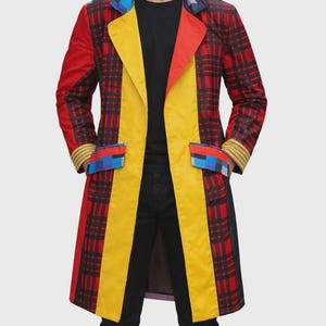 Puede incluir: Un abrigo largo y colorido con un llamativo estampado de cuadros rojos y negros. El abrigo presenta un panel central amarillo, solapas rojas y detalles azules y amarillos en los puños y bolsillos. Una pieza de moda vibrante.
