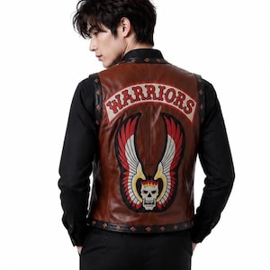 Può includere: Un gilet in pelle marrone con una grande toppa ricamata sul retro. La toppa mostra la parola WARRIORS in lettere rosse sopra un grafico di un teschio con ali. Il gilet presenta bordi neri con borchie a forma di diamante lungo i profili.