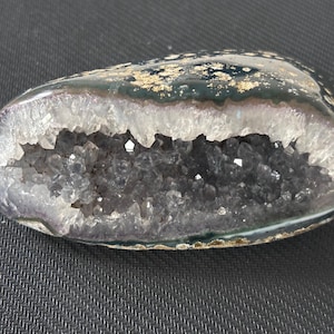 May include: An oval-shaped geode with a polished dark green and gold exterior. The interior reveals a cavity filled with sparkling, dark grey crystals. The geode's edge is lined with a band of white and purple crystals, creating a striking contrast.